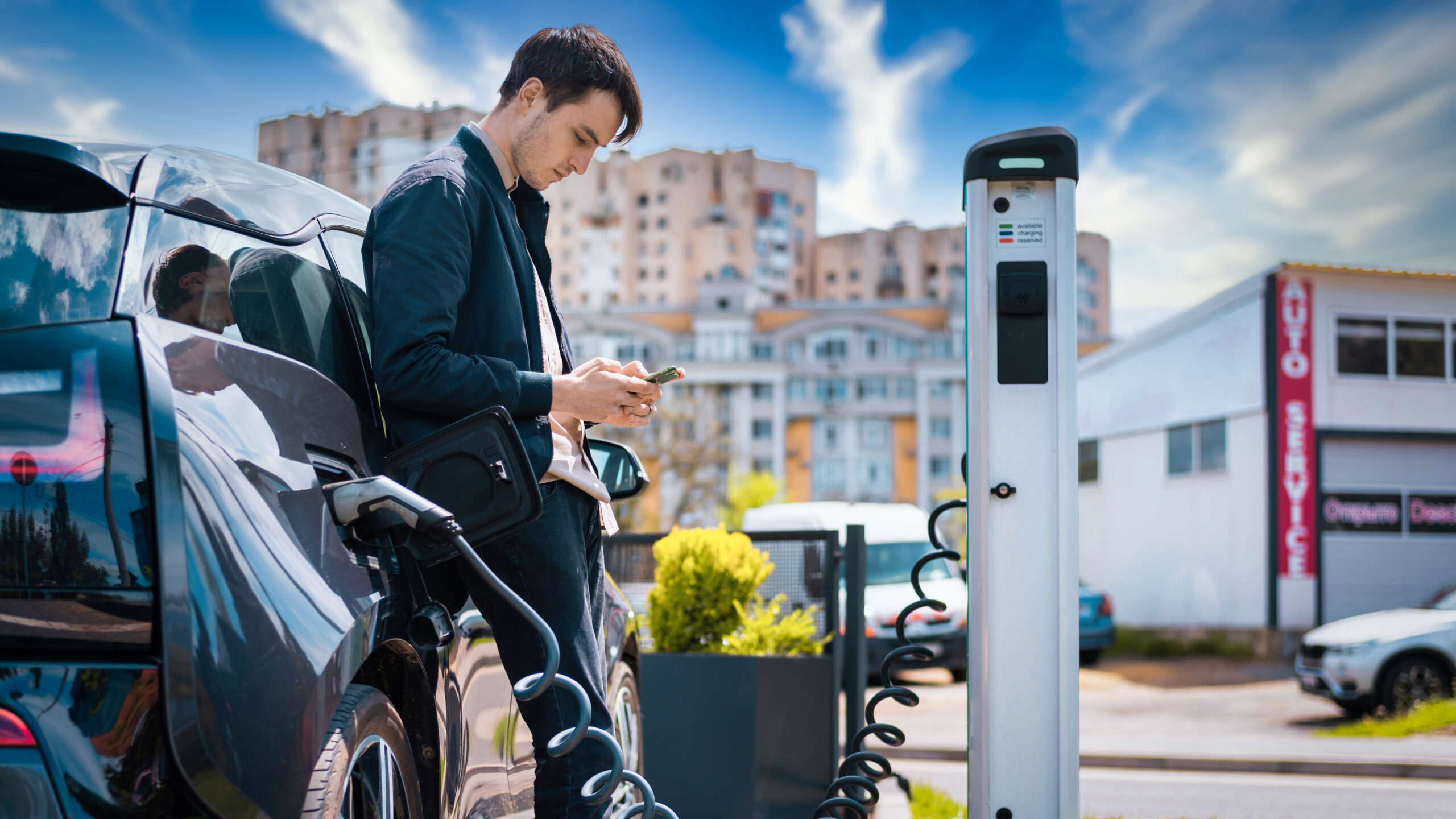 Man charging his electric car at charge station and using smartphone